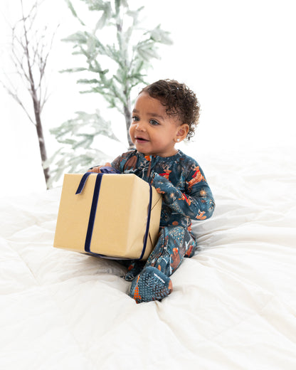 Toddler sitting on a bed holding a gift box wearing Woodland Wonder bamboo baby zip pajamas