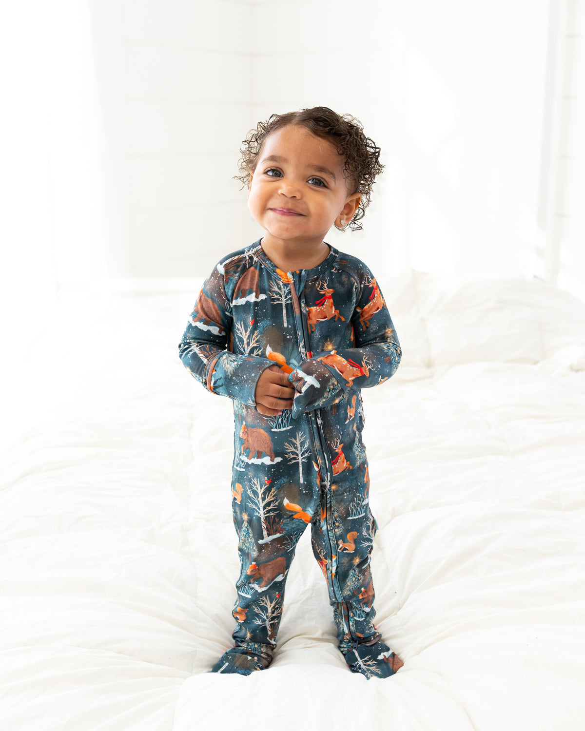 Toddler standing on a bed wearing Woodland Wonder bamboo baby zip pajamas in deep slate-blue Christmas woodland print