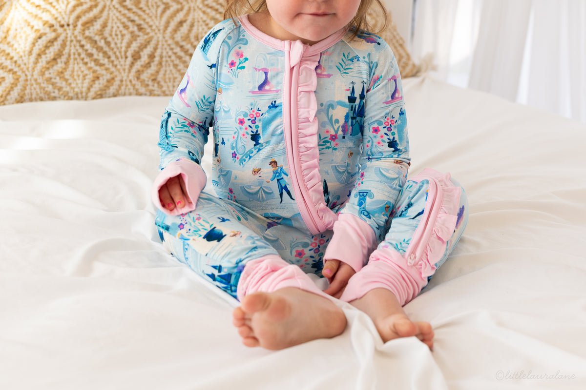 Baby Princess Pajama – The Wish Collection – on-model, toddler sitting cross-legged on bed in blue castle-print zip pajama