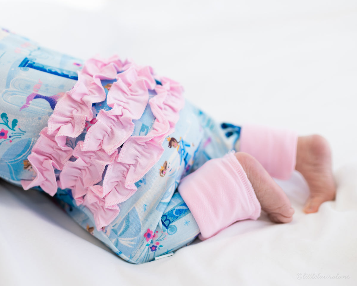 Baby Princess Pajama – The Wish Collection – on-model close-up of baby in blue castle-print zip pajama with pink ruffle