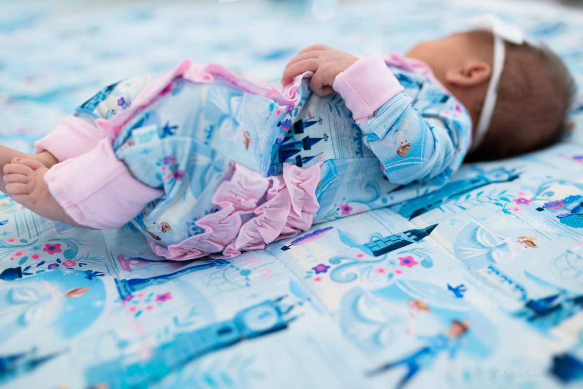Baby Princess Pajama – The Wish Collection – on-model, newborn lying on blue castle-print blanket in zip pajama