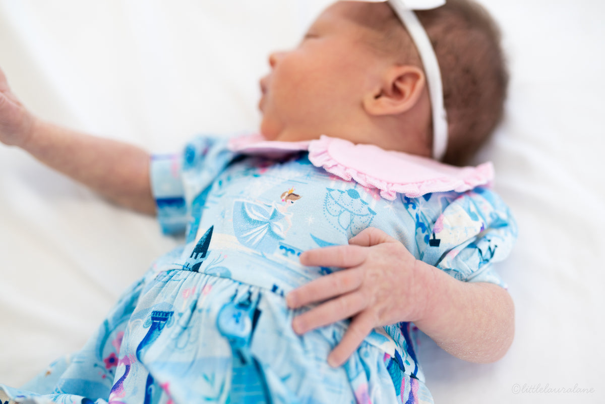 Baby Princess Dress – The Wish Collection – on-model, sleeping newborn in blue castle-print dress with pink ruffled collar